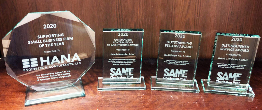 Four clear glass trophies on a brown table - a large octagonal one for Hana's Supporting Small Business Firm of the Year award, and smaller rectangular awards for other firms.